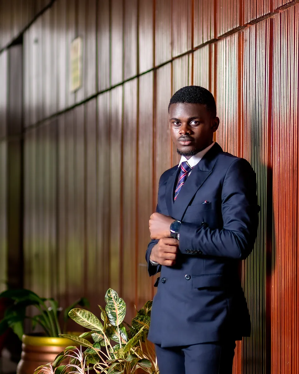 Nicholas Osei Kofi Asare in a navy double-breasted suit, standing by a wooden wall